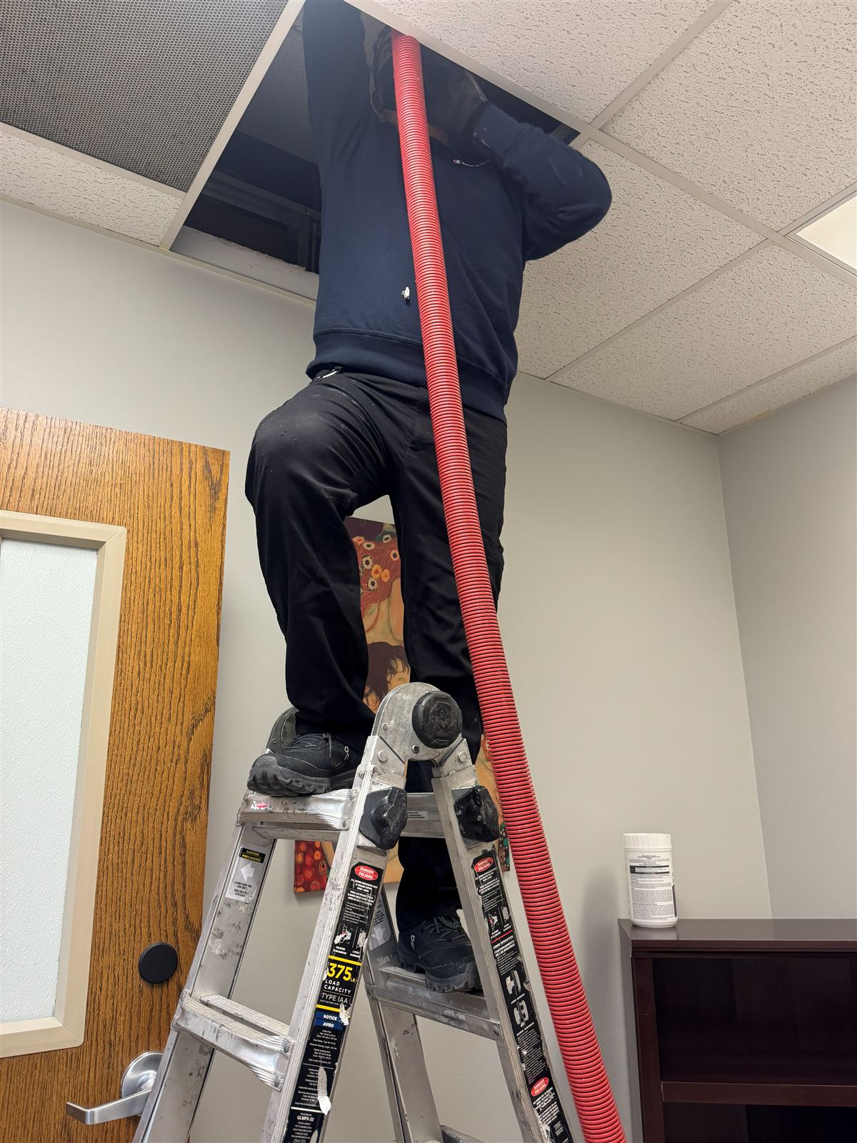 Technician on a ladder cleaning commercial air ducts through ceiling tiles in Dallas, TX